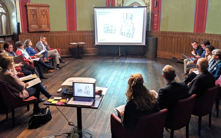 mehrere Personen sitzen in einem Kreis und diskutieren miteinander, im Hintergrund ist eine Leinwand zu sehen, auf die ein Beamer die Grundrisse des neuen Forums projeziert