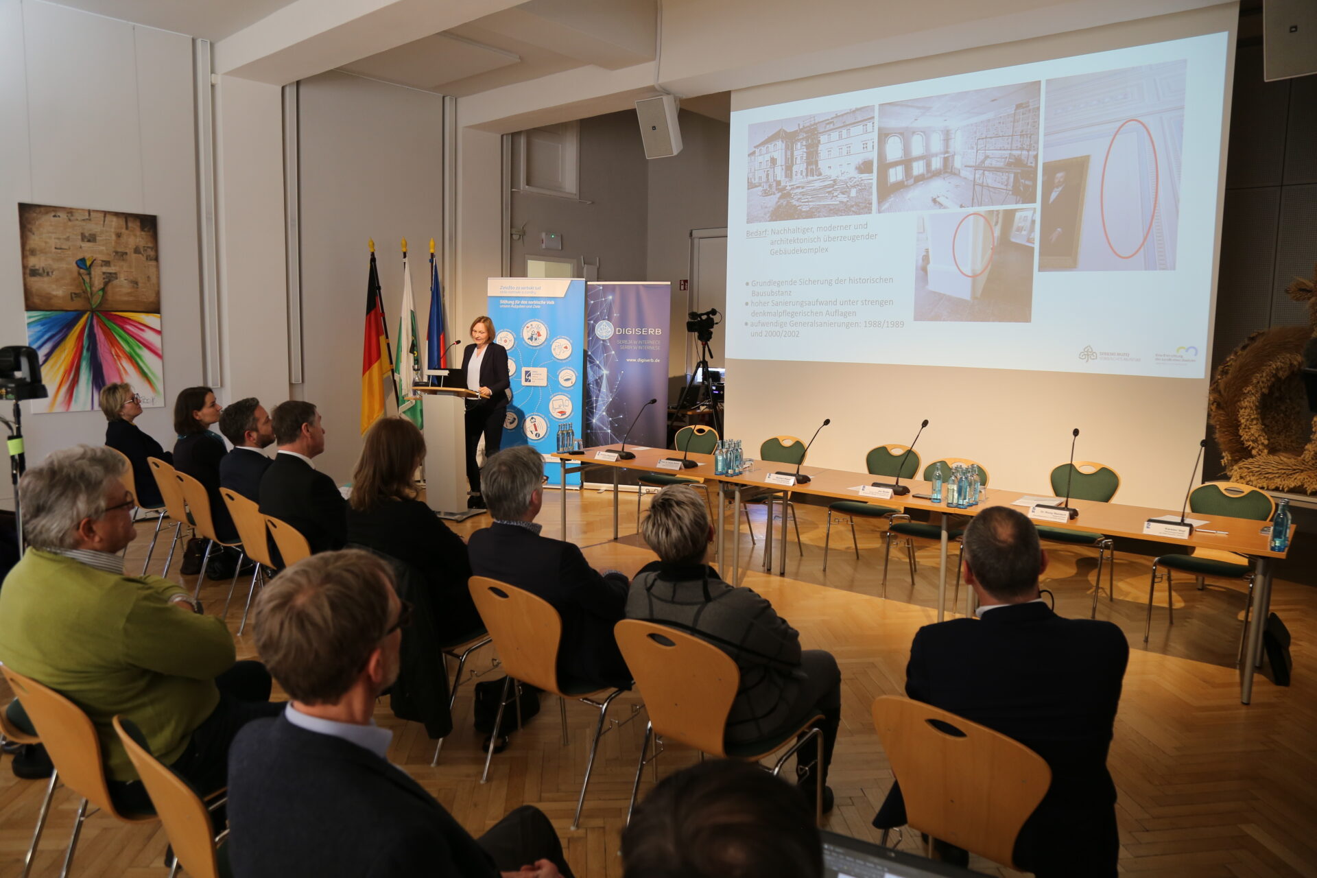 eine Frau, die einem Publikum etwas vorträgt, im Hintergrund ist die Präsentation zu sehen