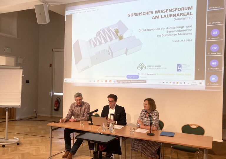 drei Personen sitzen an einem Tisch vor einer Leinwand
