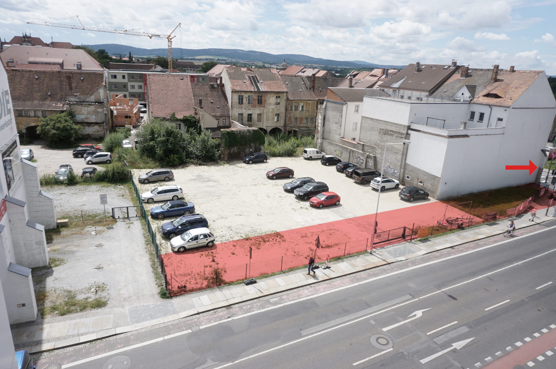 Foto mit Blick auf das zu bebauende Gelände, im Vordergrund ist ein großer Parkplatz, im Hintergrund sind mehrere Häuser, die teils sehr verfallen sind