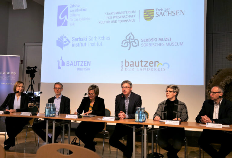 wjacore wosoby sedźa před sćěnowinu, na kotrejž pokazaja so loga wobdźělenych partnerow projekta