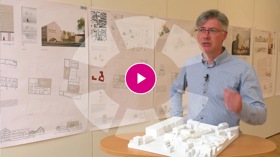 ein Mann steht vor einem Architekturmodell, im Hintergrund hängen die Pläne der Architekten