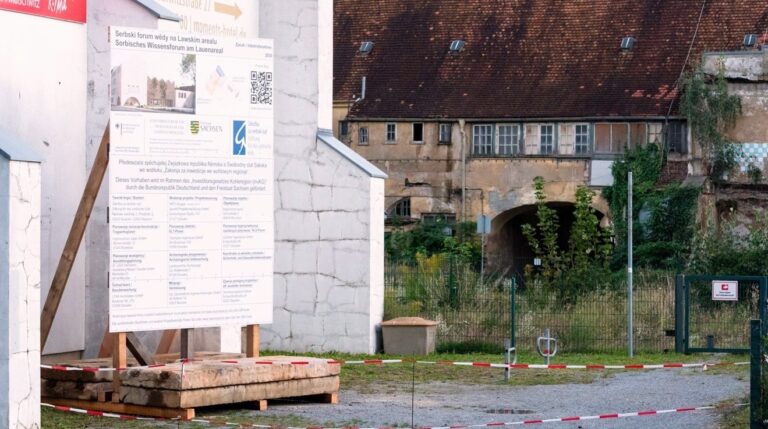 Bauschild vor einem Haus, im Hintergrund ein verfallenes Gebäude