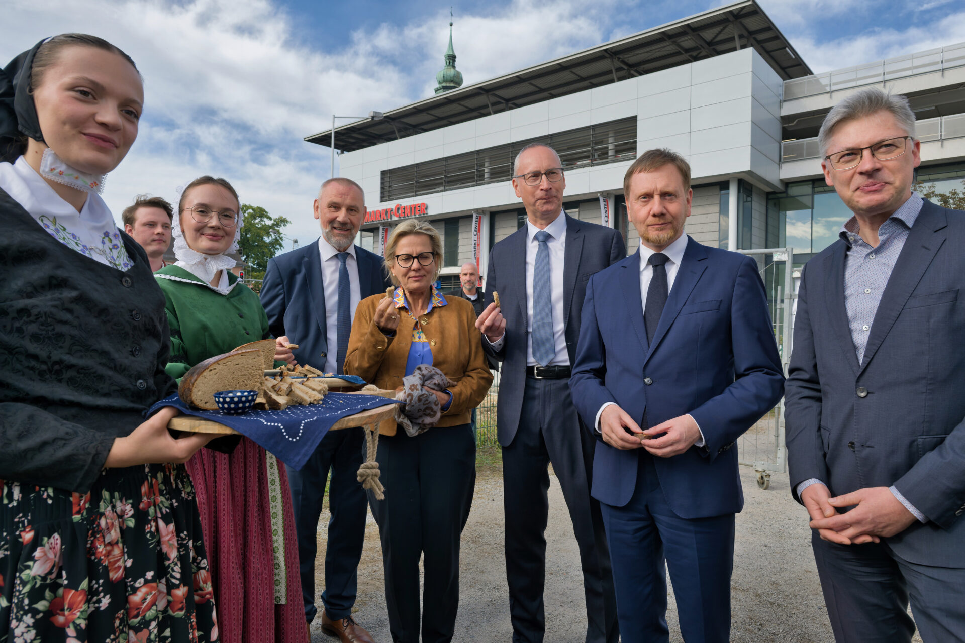 mehrere Personen stehen nebeneinander und essen ein Stück Brot, daneben zwei junge Frauen in sorbischer Tracht